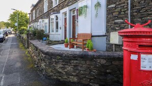 A stone wall with a bench and potted plants, a red post box, and a row of houses with white doors and windows. - Neland View (Windermere)
