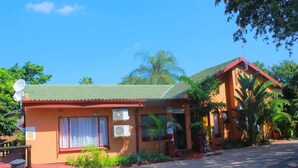 Um edifício de dois andares com telhado vermelho e paredes bege, com área de recepção, sinalização, aparelhos de ar condicionado e um pequeno jardim com plantas.