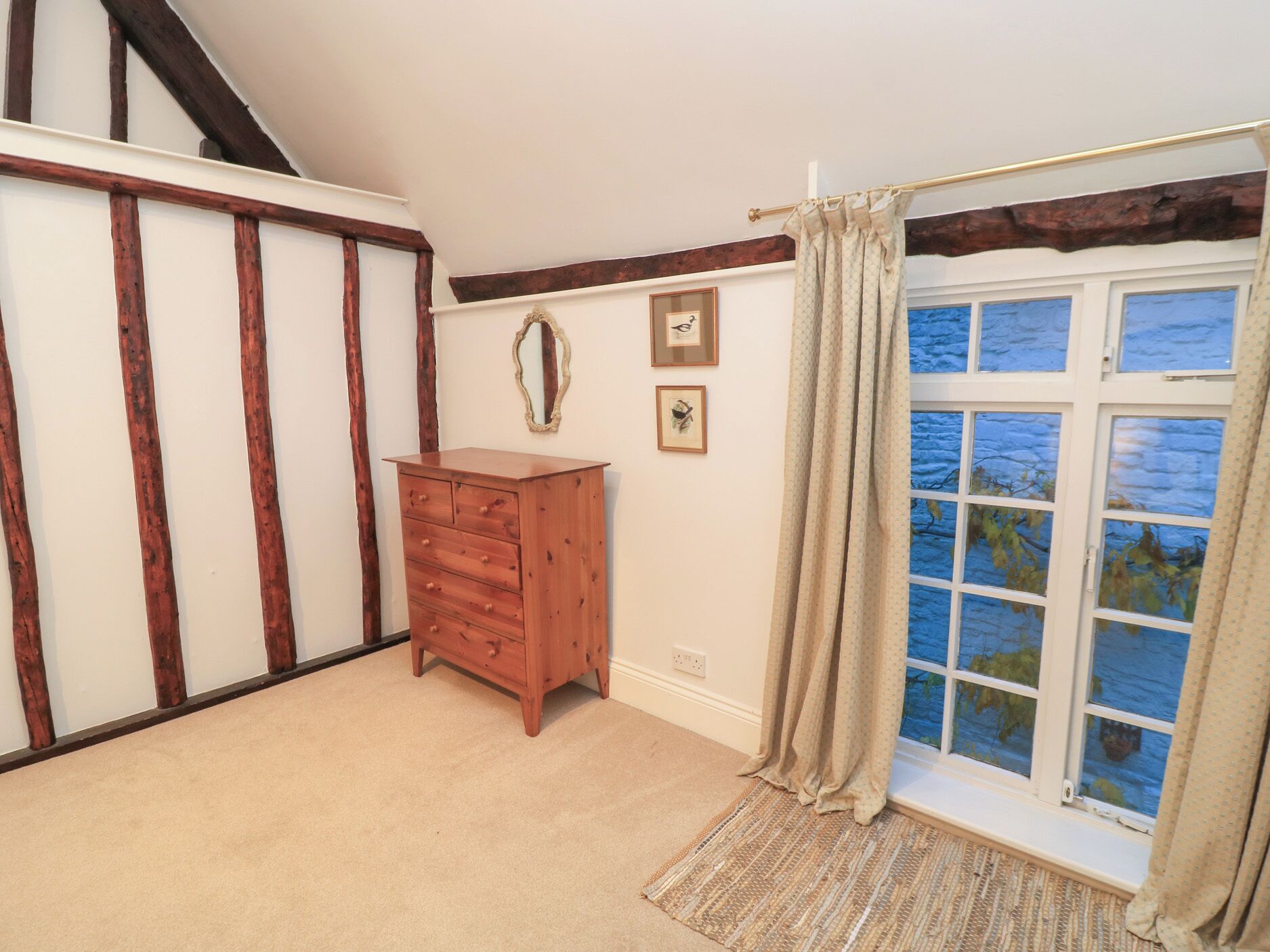 A room with exposed wooden beams, a wooden dresser, a window with a view, and a mirror on the wall.