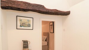 Een gang met een rustiek houten balkenplafond, een ingelijste foto aan de muur, een klein plankje met een waterkoker, en een deur die leidt naar een slaapkamer.