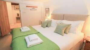 Een net opgemaakt bed met wit- en groen beddengoed, een houten nachtkastje en een wandkapstok.