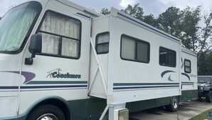 Een witte en groene camper, merk 'Couchmen' en 'MIRADA' zichtbaar, met een schuifdeur, ramen en een klein aanhangend hulpkastenbakje.