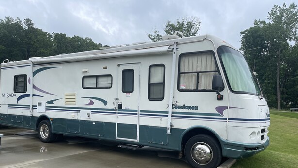 Een witte camper met 'Couchmen'-branding, geparkeerd op een betonnen oppervlak, met gras en bomen op de achtergrond.