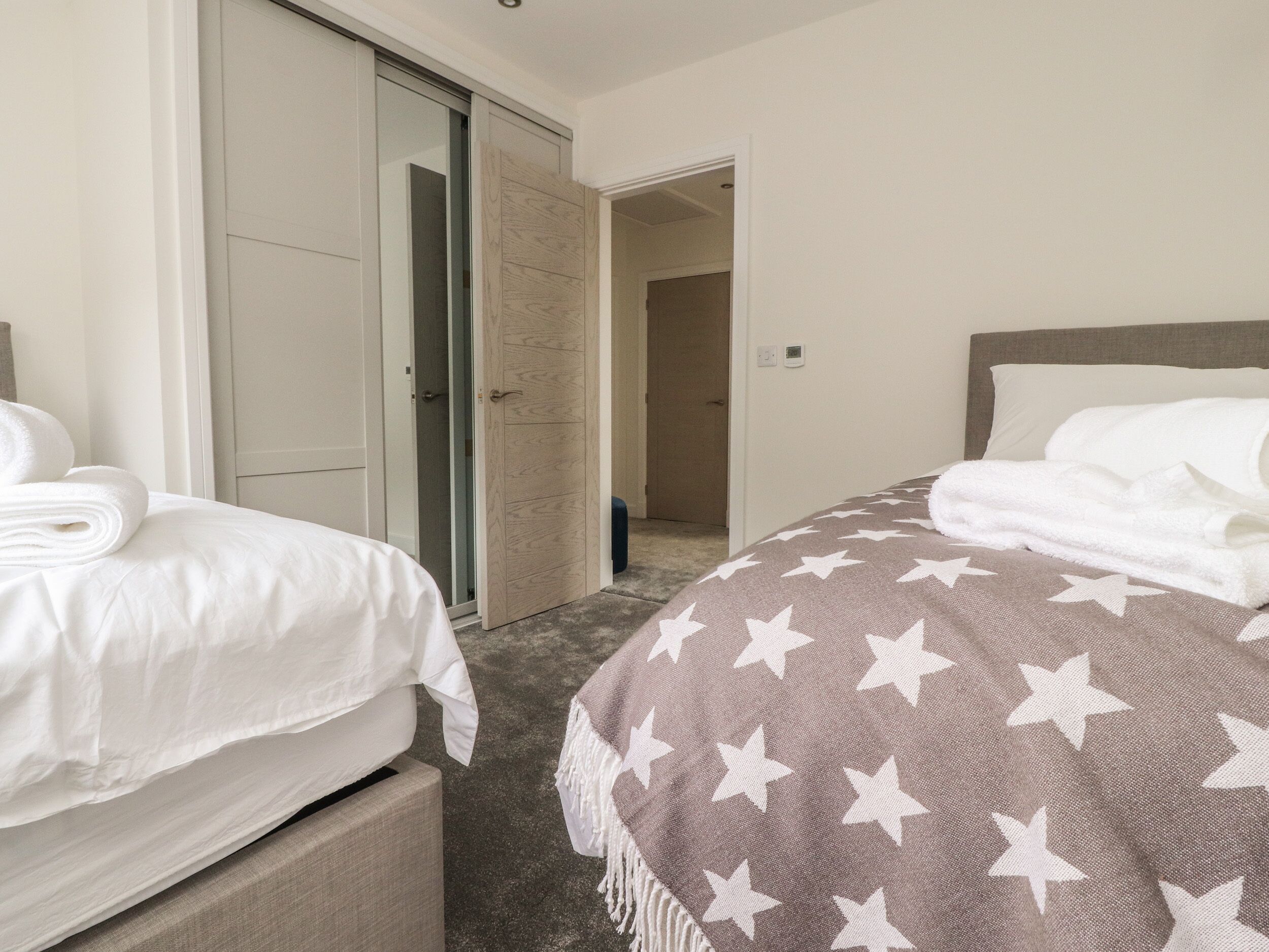 Une chambre d’hôtel avec deux lits, l’un recouvert d’une courtepointe blanche et l’autre d’une courtepointe marron ornée d’étoiles blanches. La chambre est dotée d’une porte en bois et d’un petit tabouret bleu.