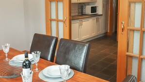 Un comedor con una mesa de madera dispuesta para dos personas, una botella de vino y dos copas de vino. Se ven armarios blancos y un microondas al fondo.