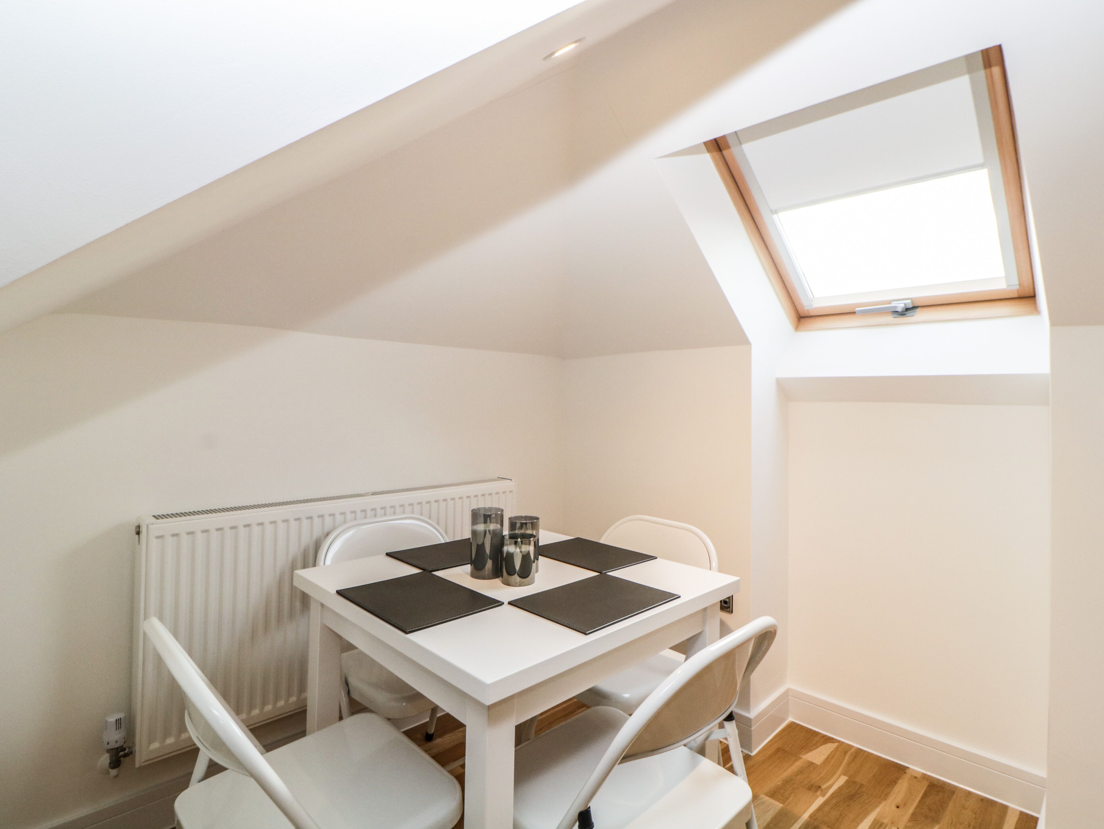 Un petit coin repas, de couleur blanche, avec une table et quatre chaises, un radiateur et une lucarne.