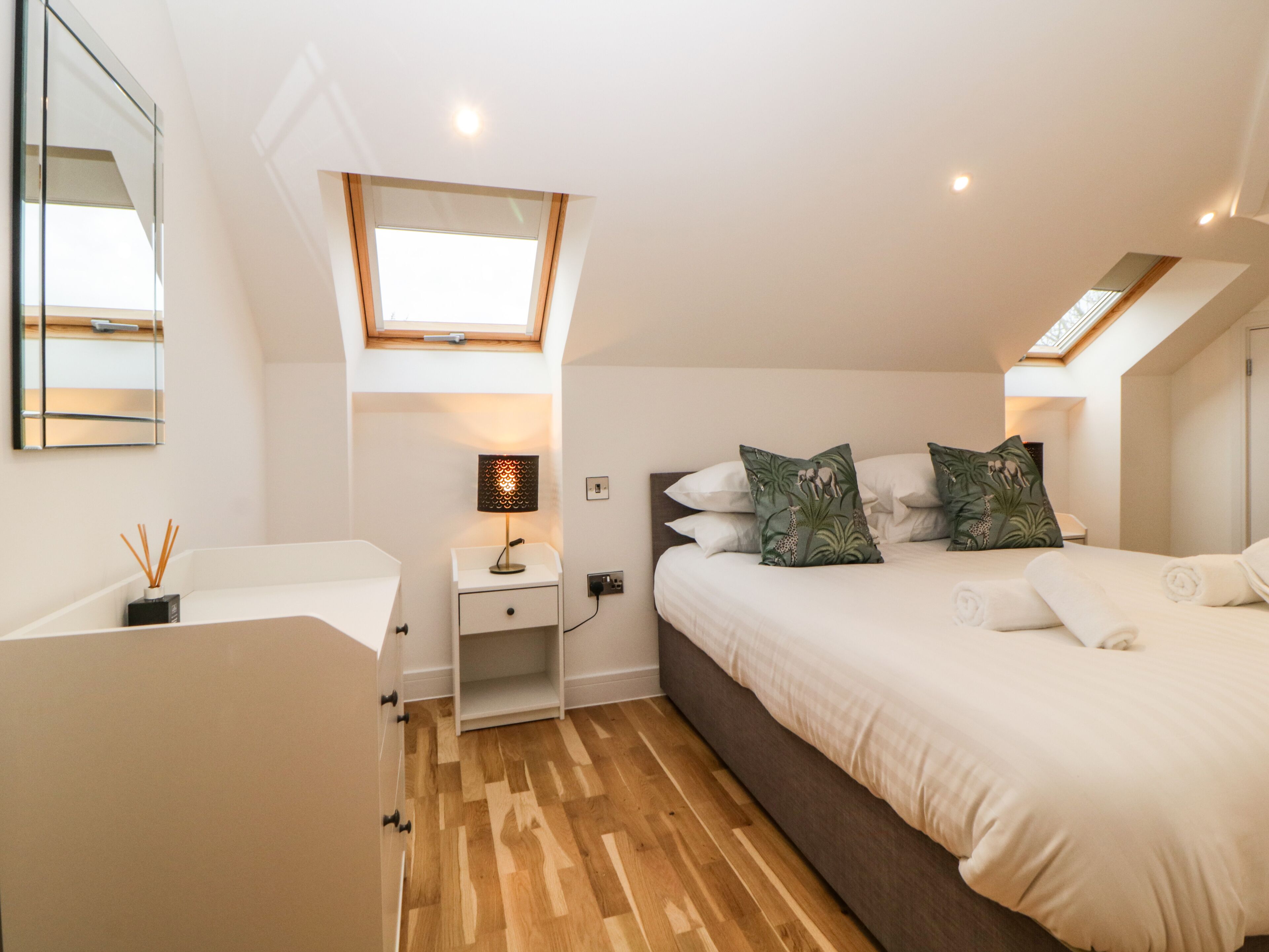 A bedroom with a bed, a nightstand, a lamp, a mirror, and a skylight.