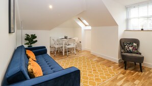 Ein modernes Wohnzimmer mit einem blauen Sofa, einem Essbereich mit weißem Tisch und Stühlen sowie einem kleinen Sessel mit einem Blumenkissen.