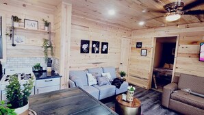 Un salon confortable avec un canapé, une table basse et un téléviseur. Il y a également un coin cuisine équipé d’un évier et d’une cuisinière. La pièce a des murs en bois et un ventilateur de plafond.
