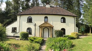 Une maison à deux étages, avec un toit recouvert de tuiles, une entrée centrale surmontée d’un arc et un jardin où poussent diverses plantes et fleurs.