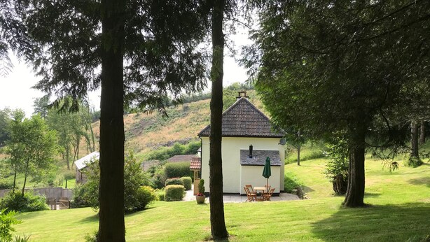 Une petite maison avec un toit en tuiles, entourée d’une végétation luxuriante et d’une pelouse bien entretenue.