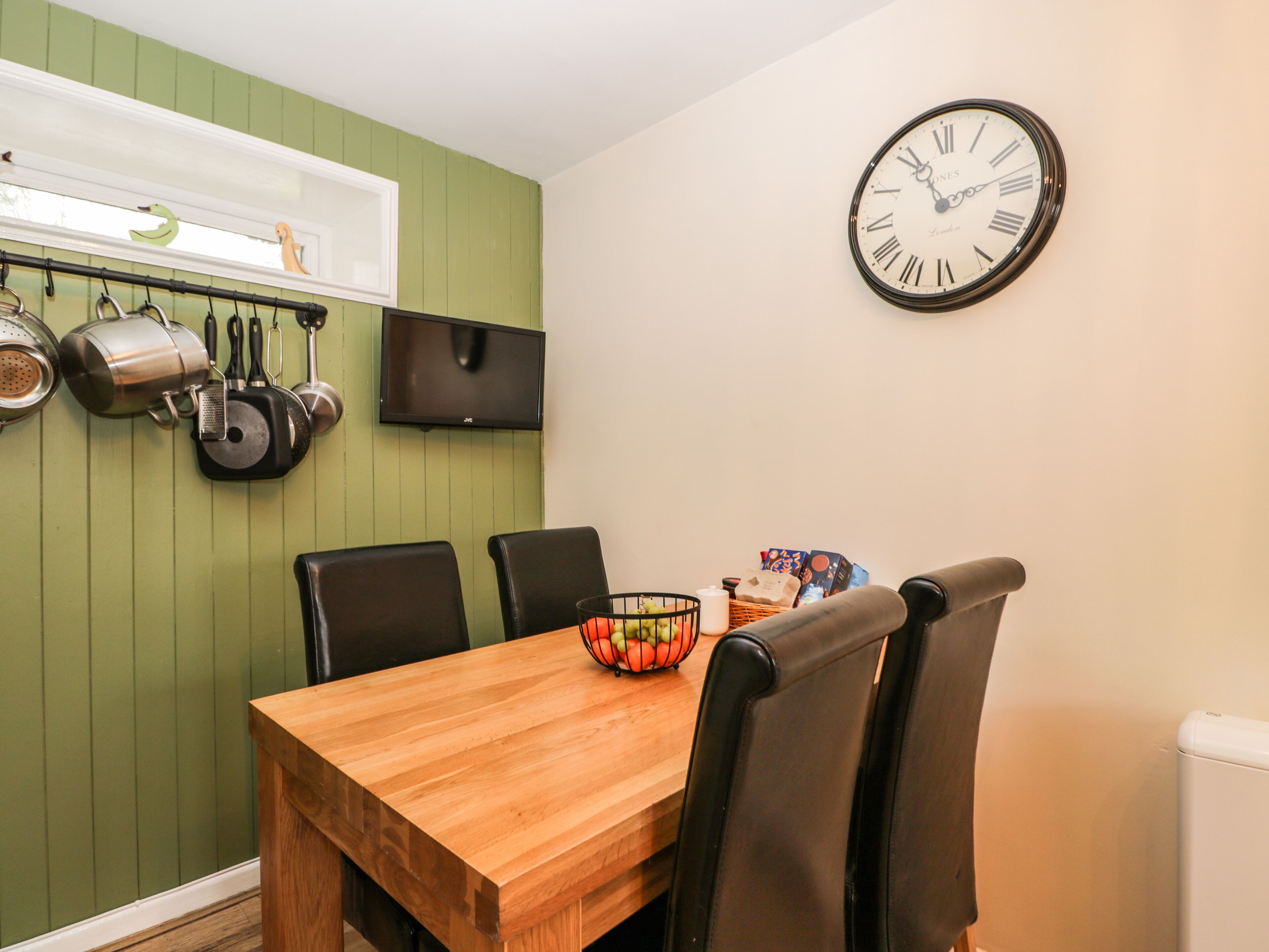 Un comedor con una mesa de madera, cuatro sillas, un reloj en la pared y utensilios de cocina colgando.