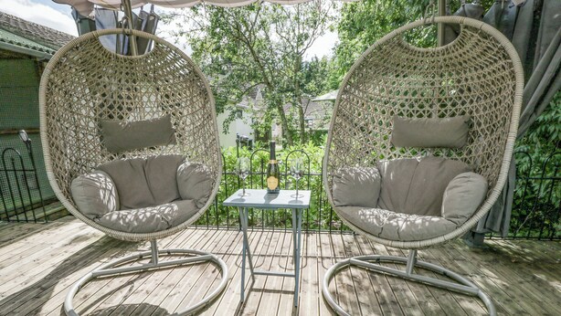 Two wicker chairs with cushions, a small table with a bottle and glass, and a wooden deck. - Orchard House Cottage (Malmesbury)