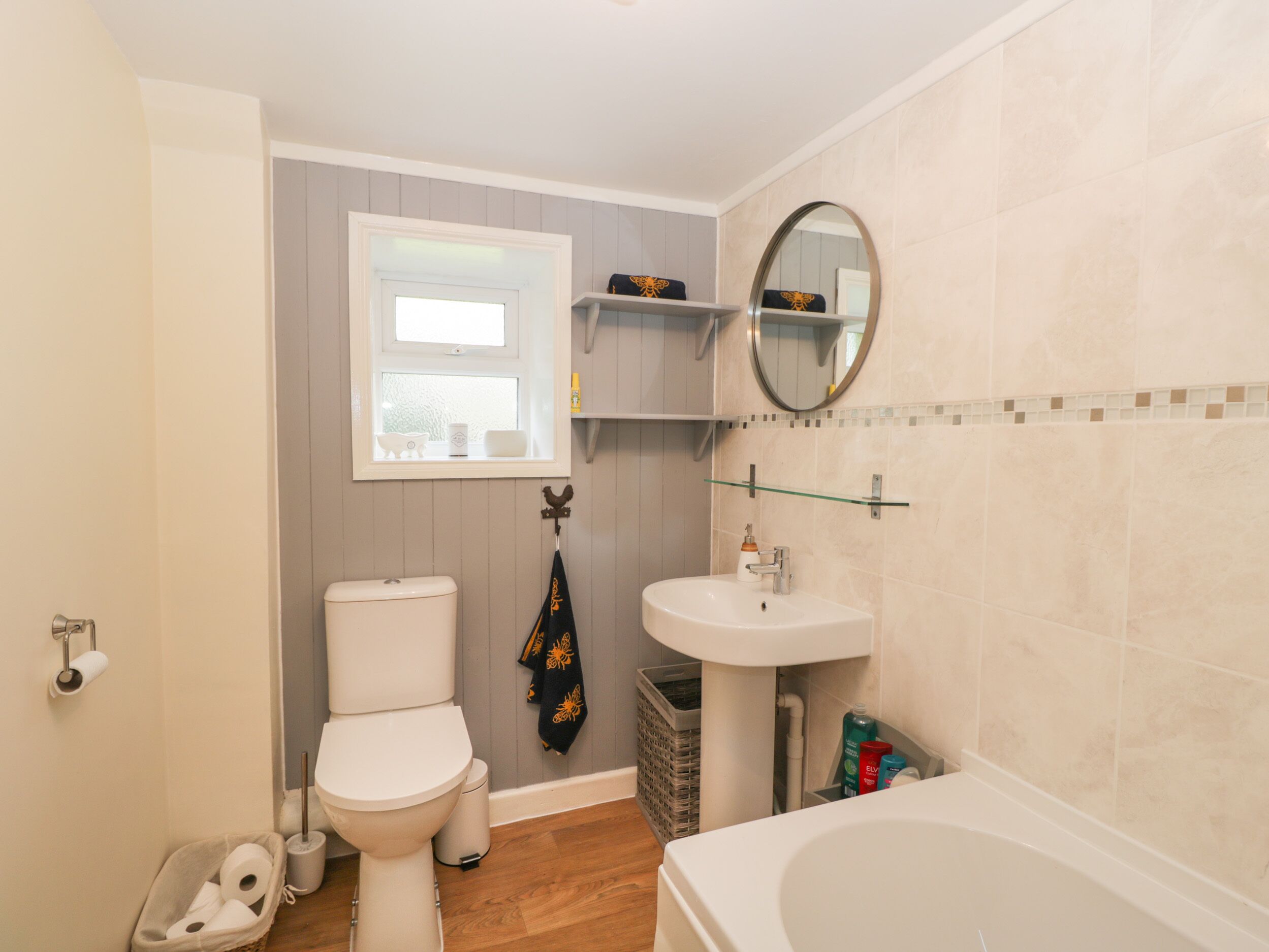 Un baño con inodoro, lavabo y bañera. Hay una ventana sobre el inodoro, un espejo sobre el lavabo y un estante encima del lavabo.