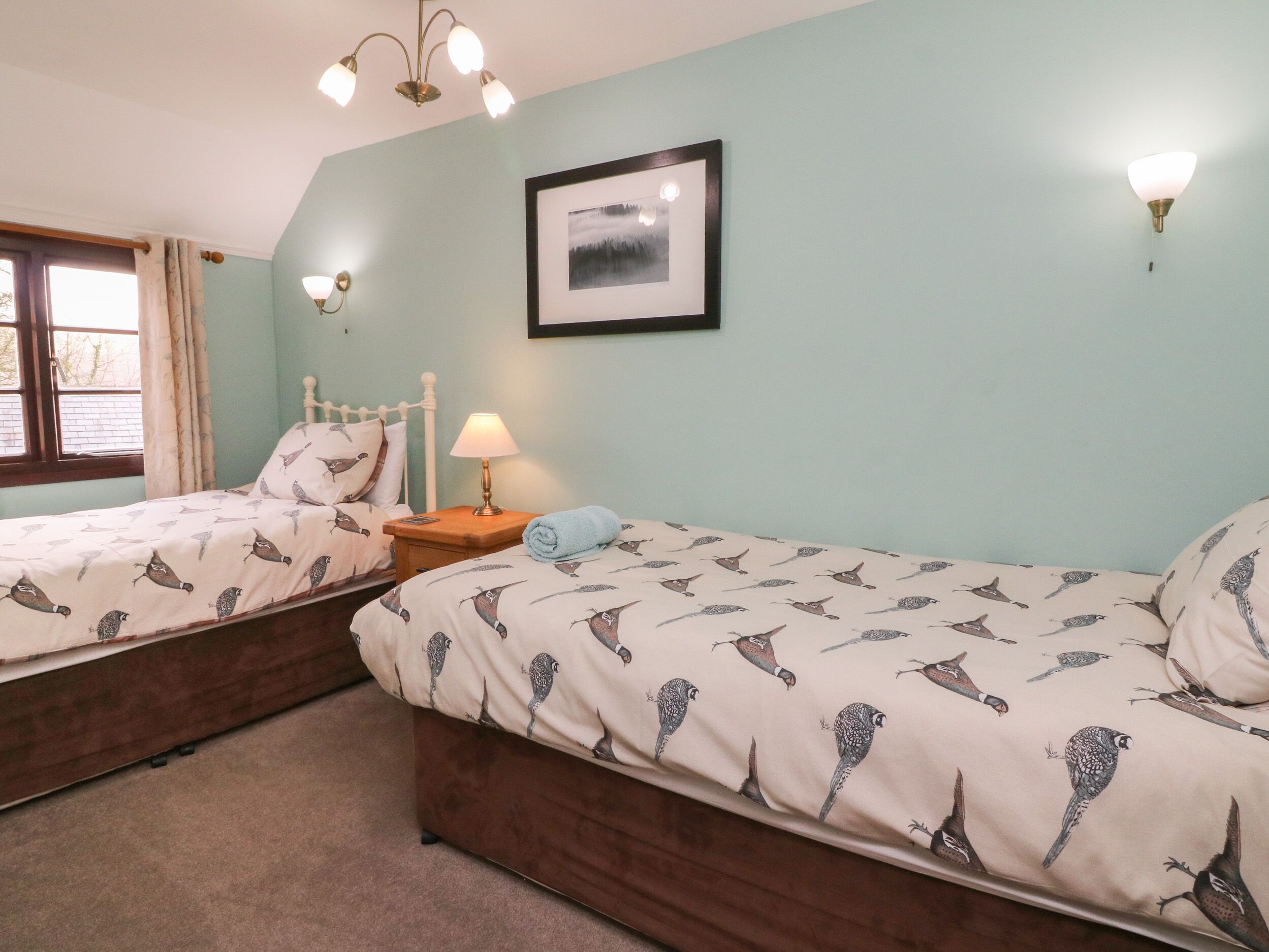 Un dormitorio con dos camas individuales, una mesita de noche con lámpara, un cuadro en la pared y una ventana con cortinas.