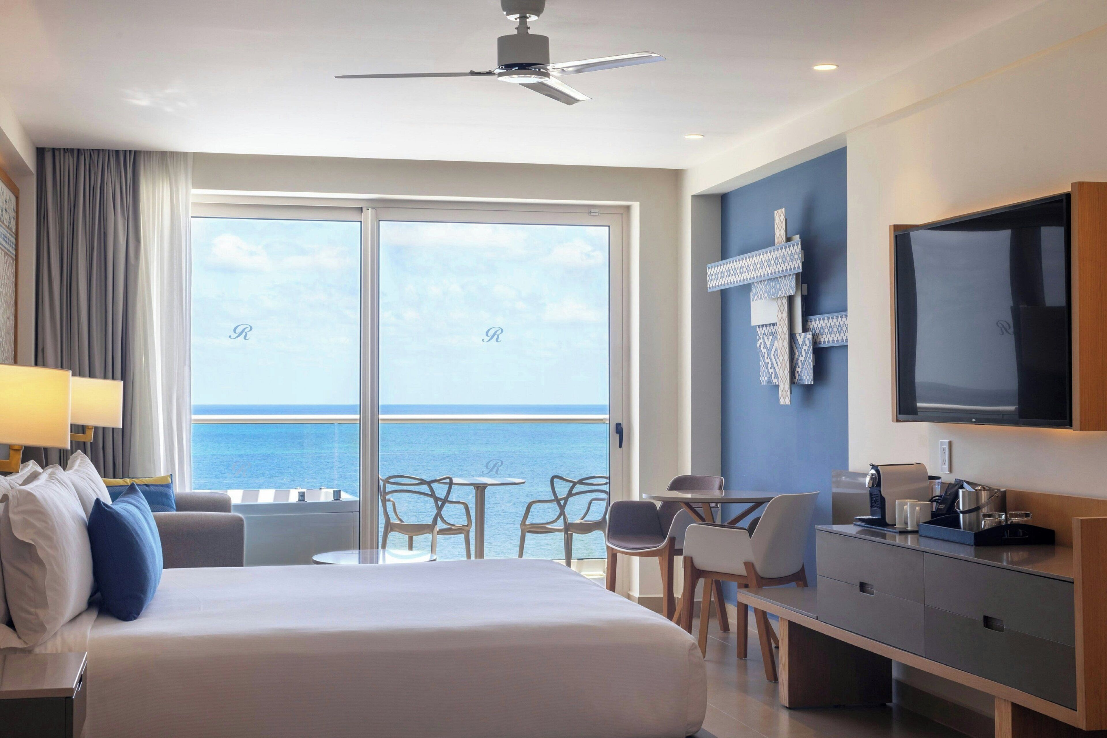 A hotel room with a bed, a desk, a TV, and a balcony with a view of the sea.