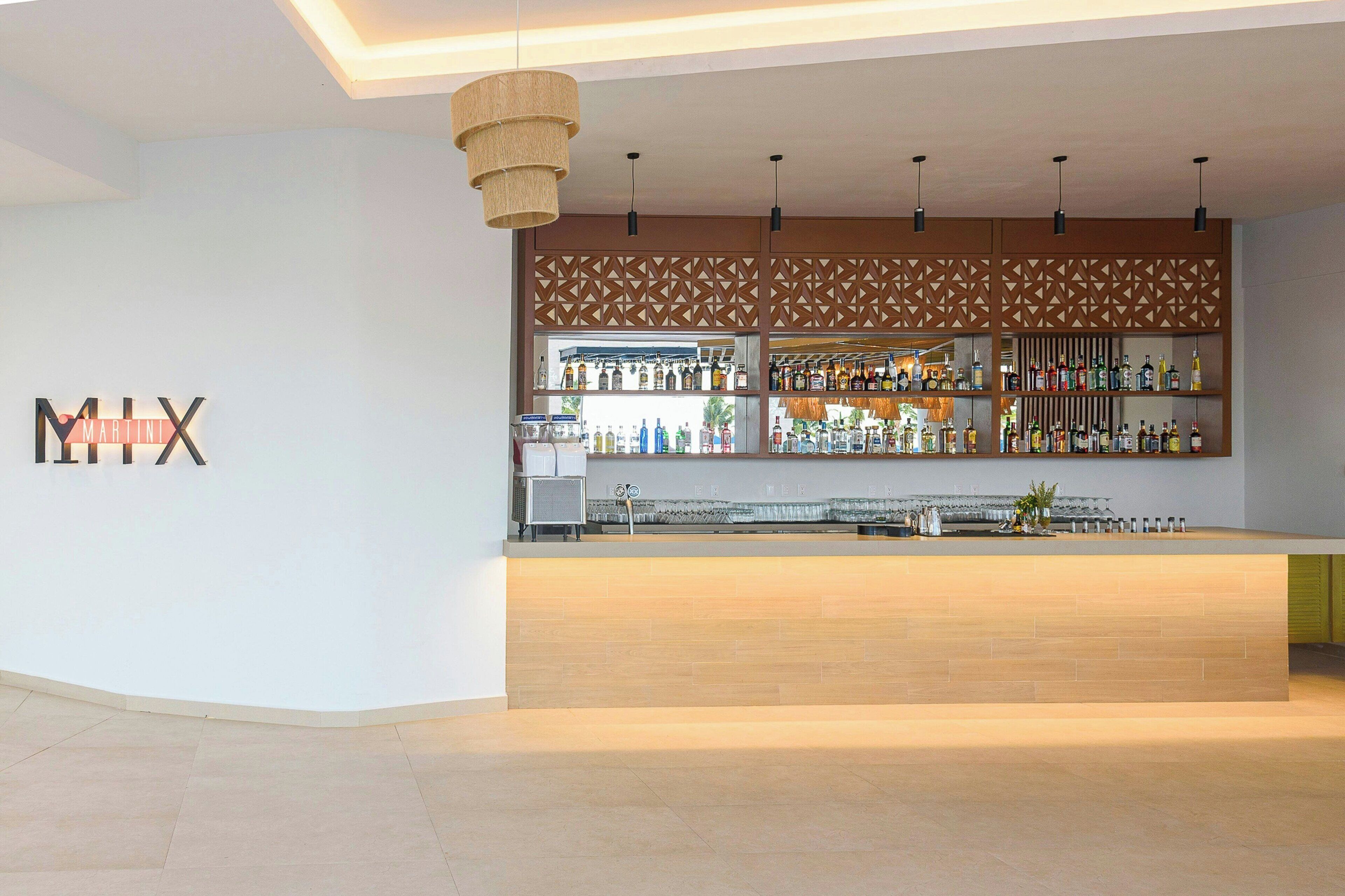Un espace bar moderne avec un comptoir en bois, une variété de bouteilles d’alcool et un mur décoratif orné de motifs géométriques.