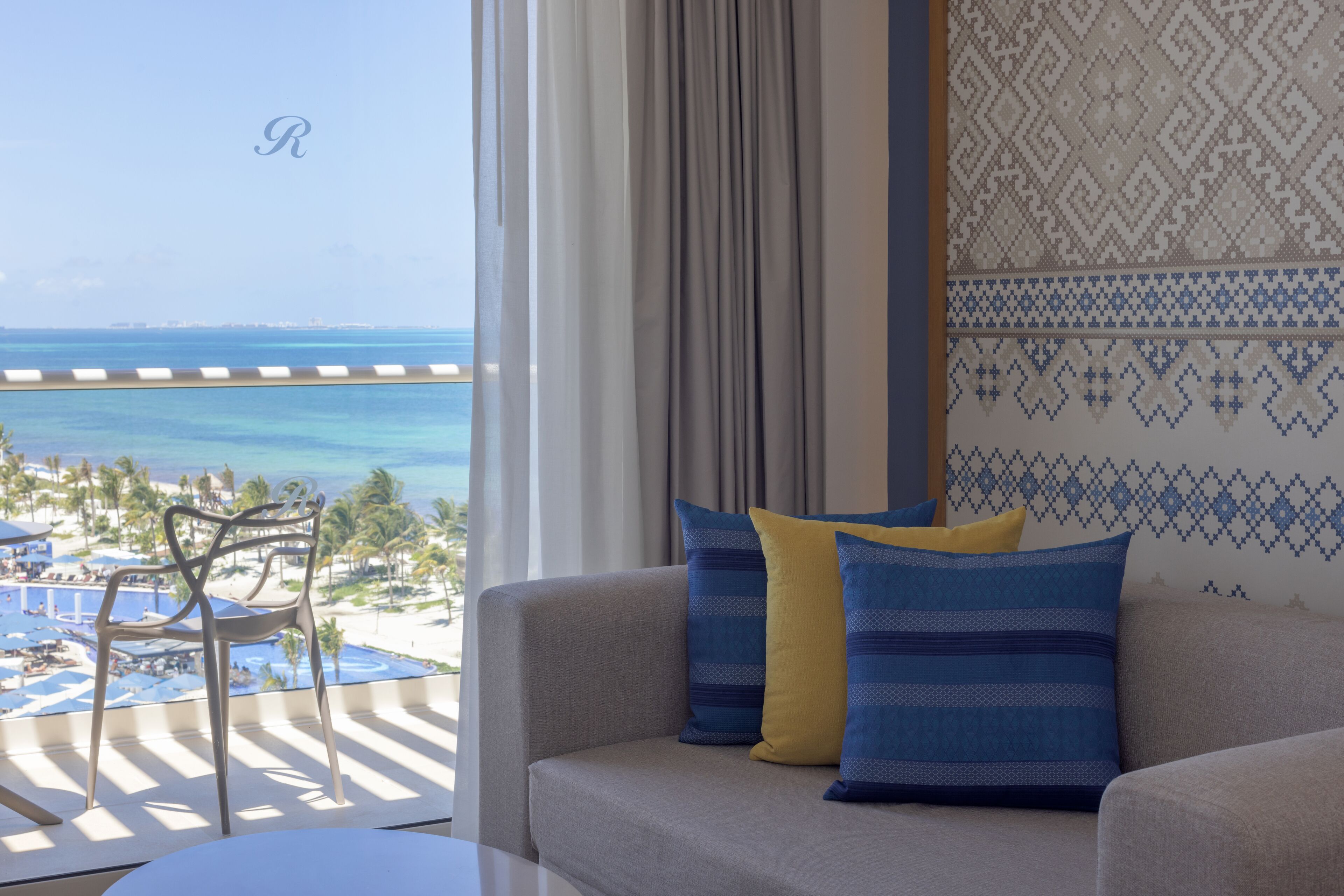 A room with a view of a beach and ocean, a sofa with patterned cushions, and a chair by the window.