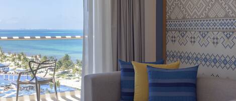 A room with a view of a beach and ocean, a sofa with patterned cushions, and a chair by the window.