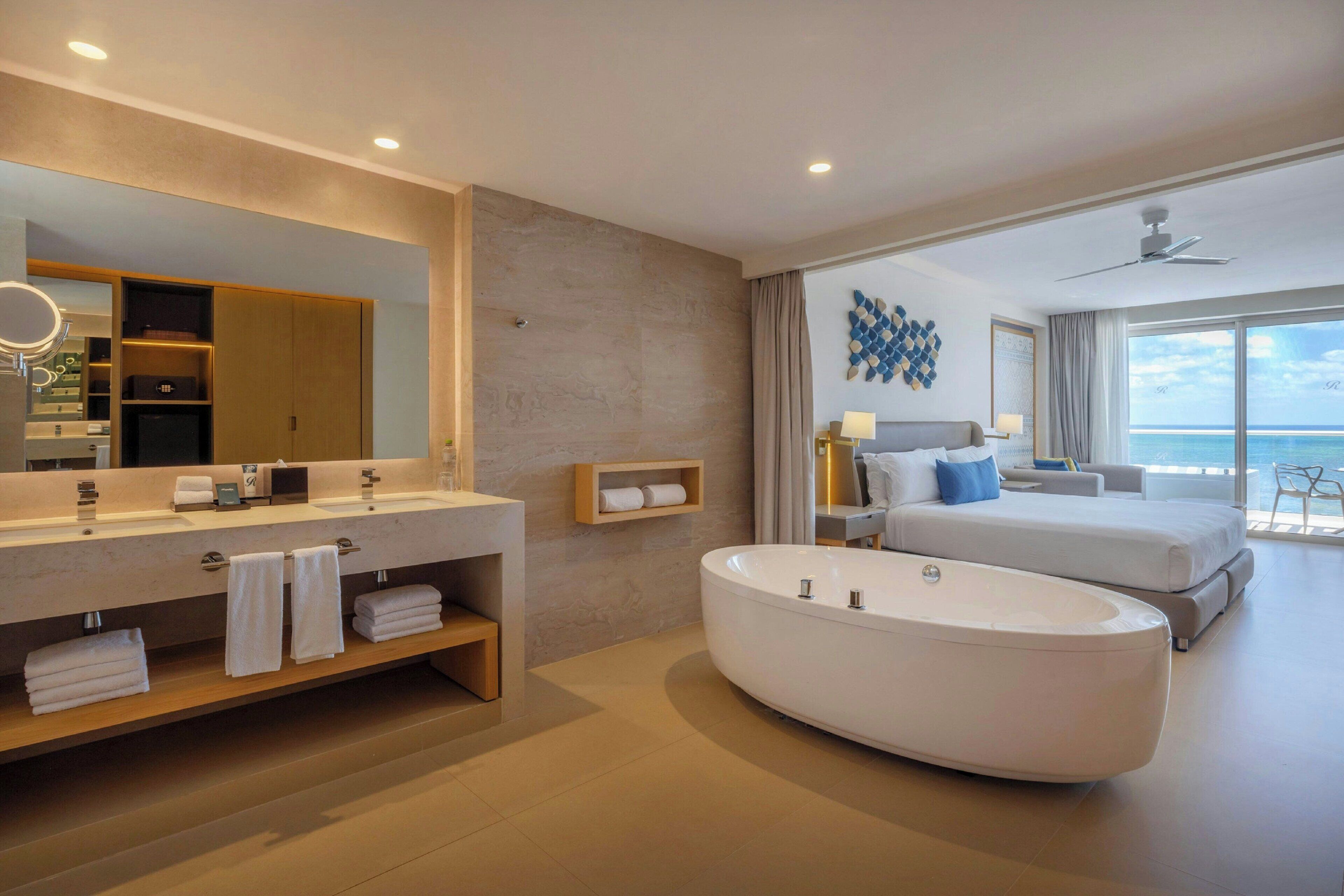 A modern hotel room with a large bathtub, a double vanity with a mirror, a bed with a view of the sea, and a ceiling fan.