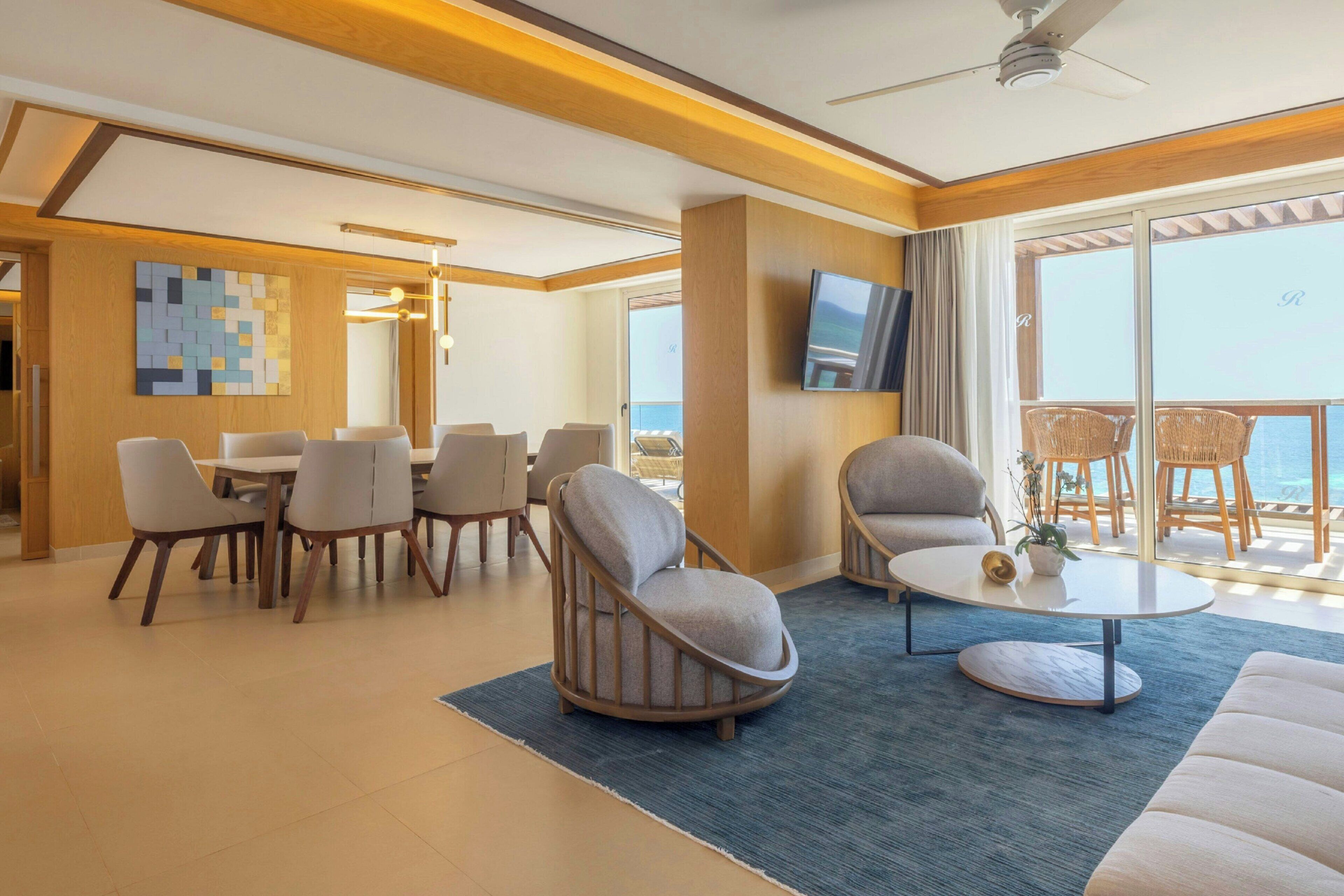 Une chambre d’hôtel moderne comprenant un coin repas, un salon avec un canapé et des fauteuils, un téléviseur à écran plat fixé au mur et un balcon aménagé avec des meubles en osier.