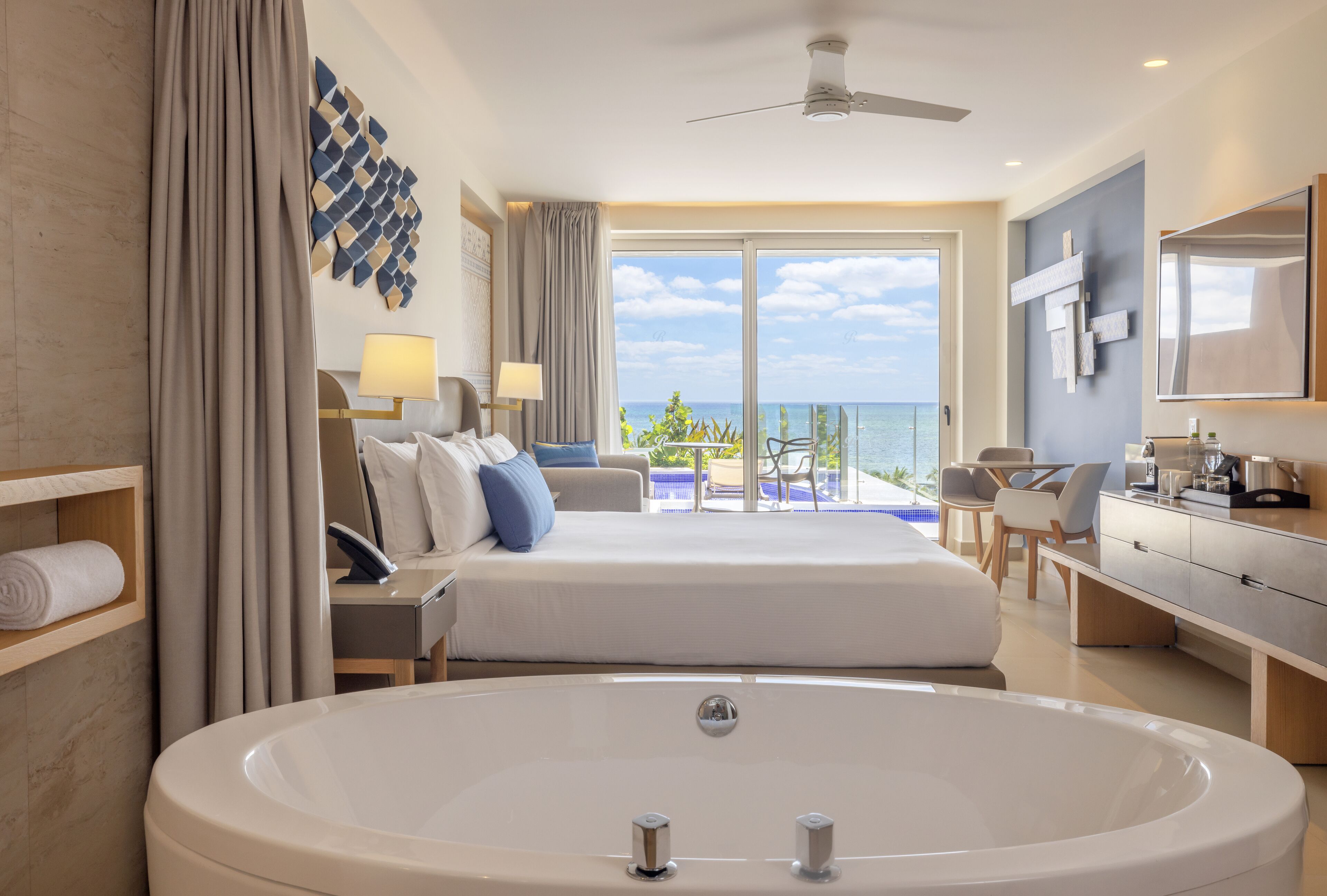 A modern hotel room with a large bathtub, a balcony with a pool view, and a desk with a chair.