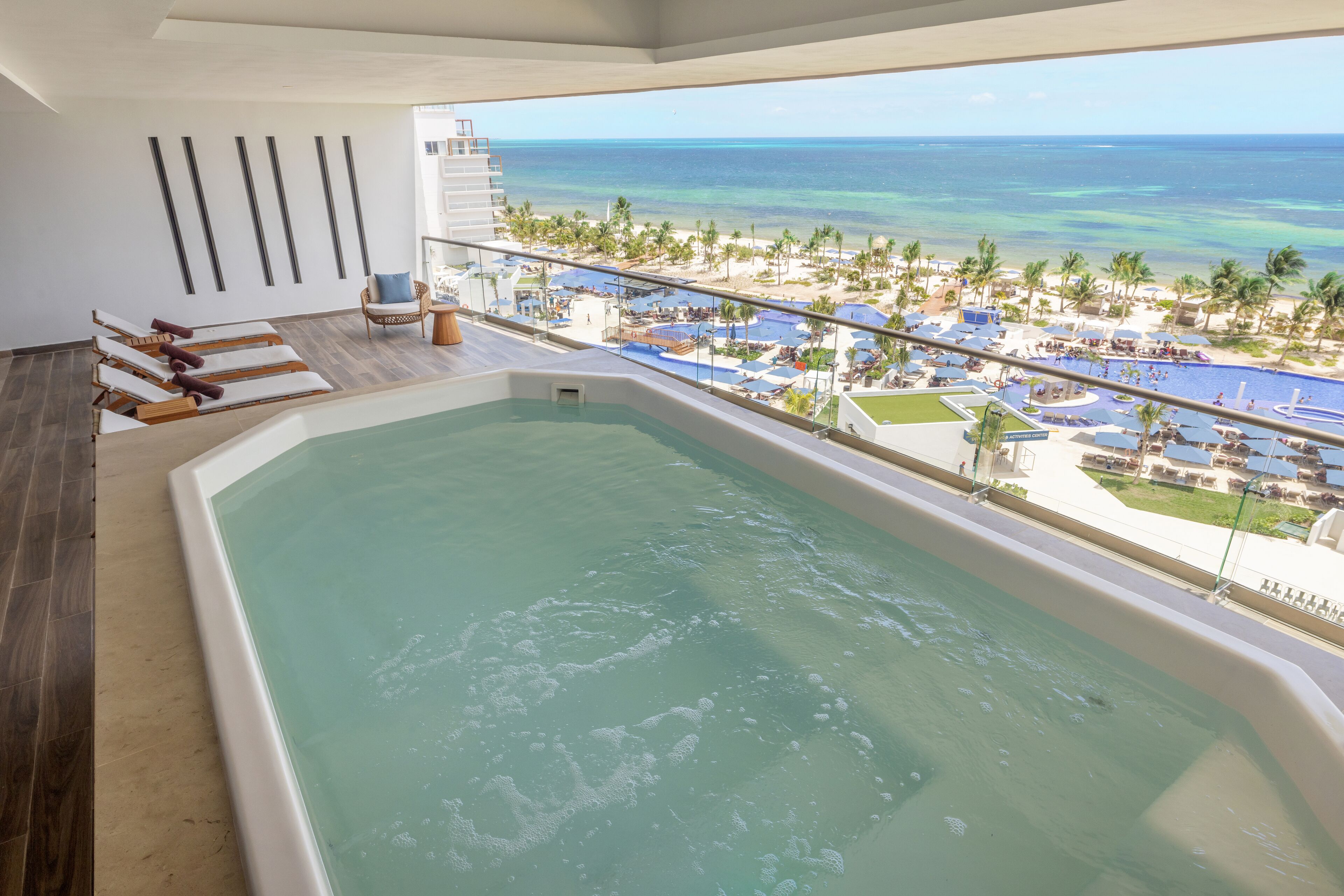 A balcony with a view of the ocean, a pool, and a lounge area with chairs and a sun lounger.
