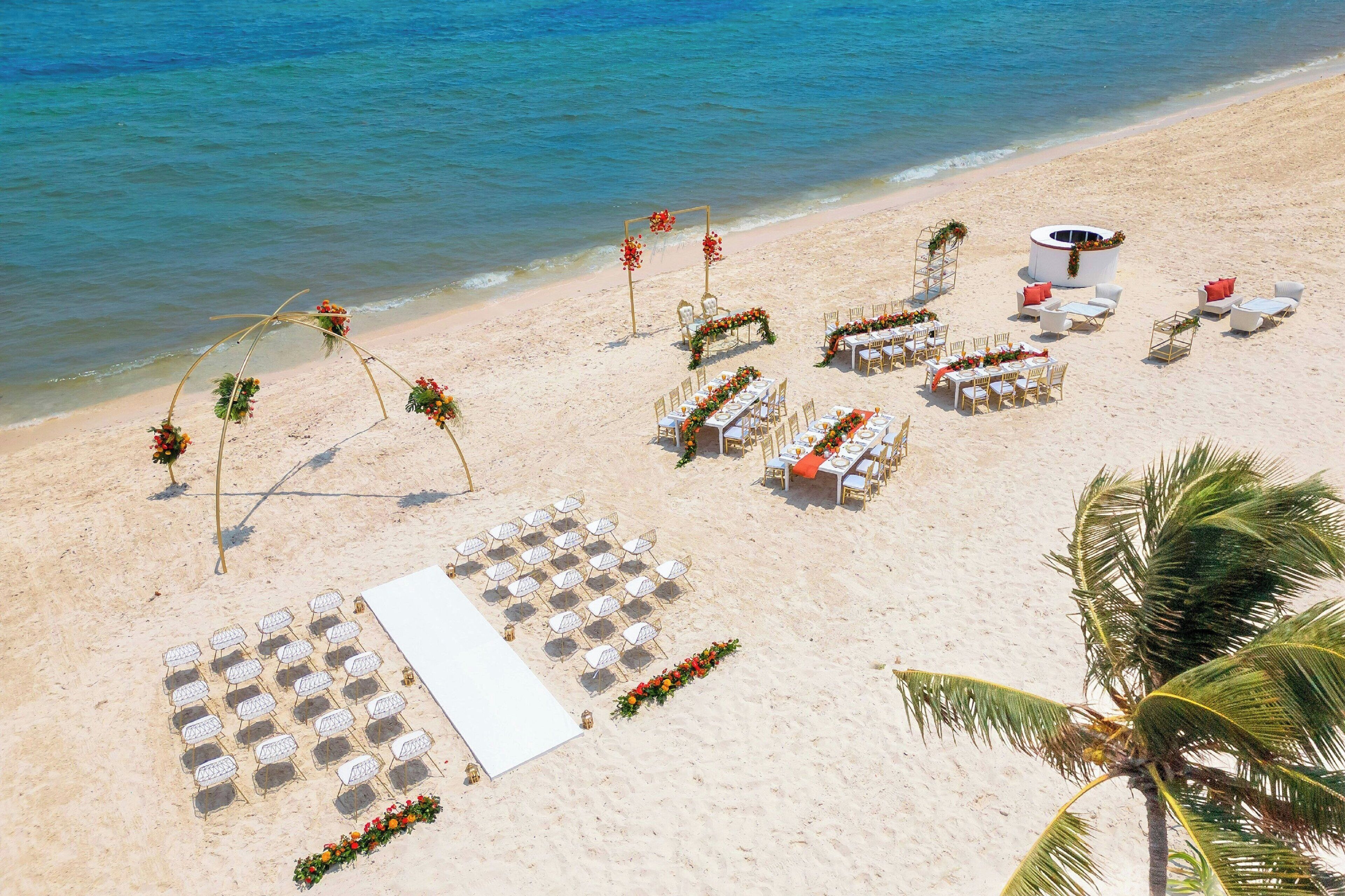 Decoración de boda en la playa que incluye un arco blanco, mesas, sillas y una pequeña estructura.