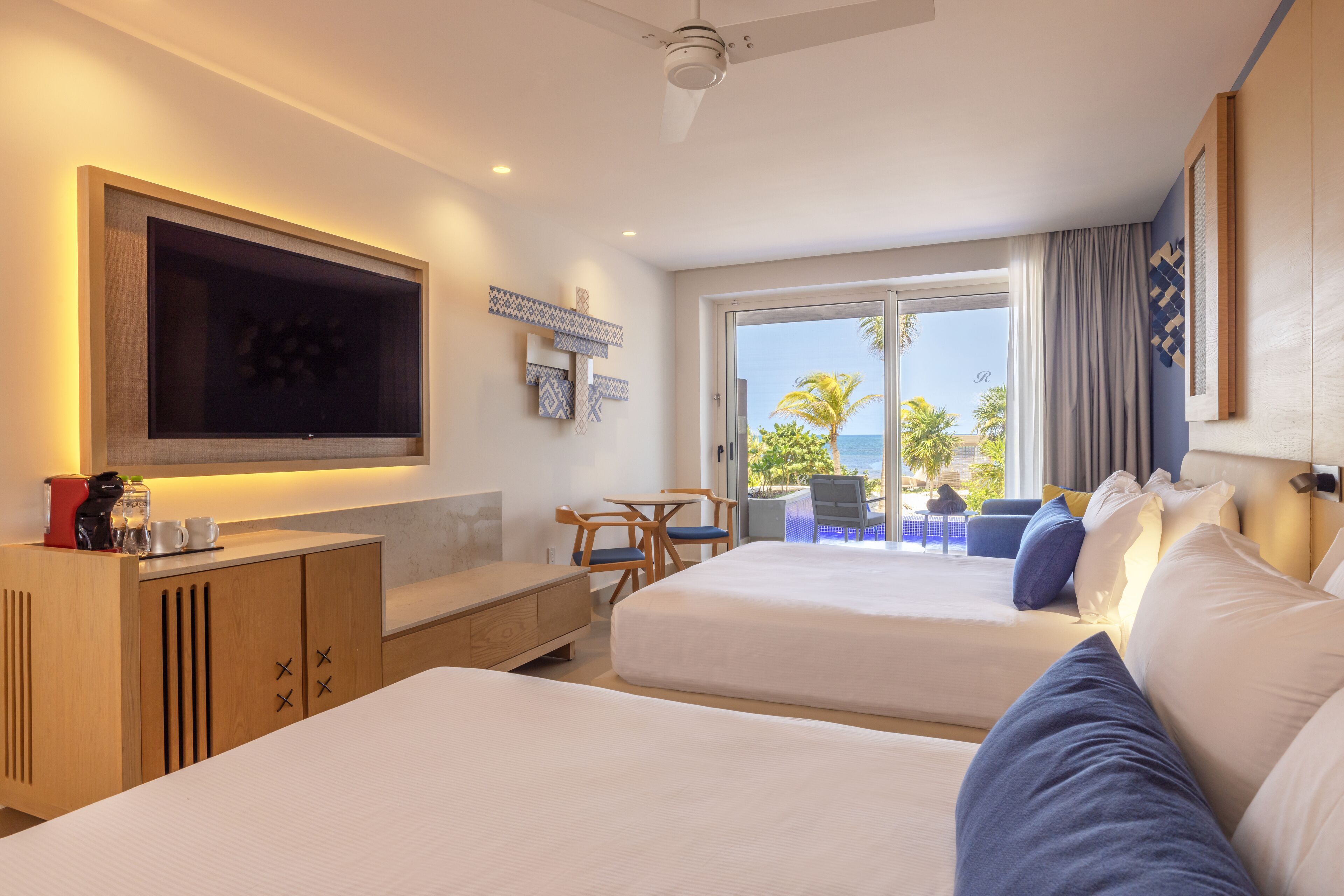 A hotel room with two beds, a TV, a desk, and a balcony with a view of the sea.
