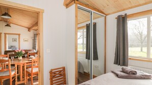 A cozy room with a wooden ceiling, a bed with a rolled towel, a dining table with chairs, and a view of the outdoors through a window.