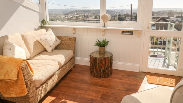Ein gemütliches Zimmer mit einem Korbsessel, einem kleinen Holztischchen mit einer Pflanze und Blick auf eine Stadtlandschaft durch große Fenster.
