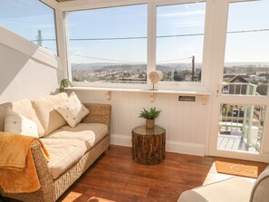 A cozy room with a wicker sofa, a small wooden side table with a plant, and a view of a cityscape through large windows. - Pebble Lodge (Paignton)