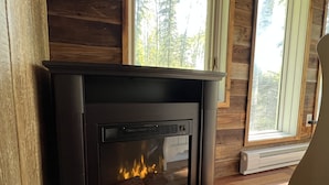 A fireplace with a visible fire, wooden walls, and a window with a view of trees. - Cozy year round cabin in the woods, walking distance to the lake (Buck Lake AB)