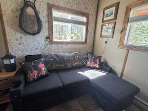 A room with a dark grey sofa, floral wallpaper, a wooden-framed window, and a wall-mounted lantern.