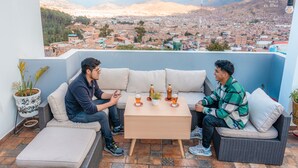 Una terraza con sofá y mesa, dos personas sentadas, y vista panorámica de la ciudad al fondo.