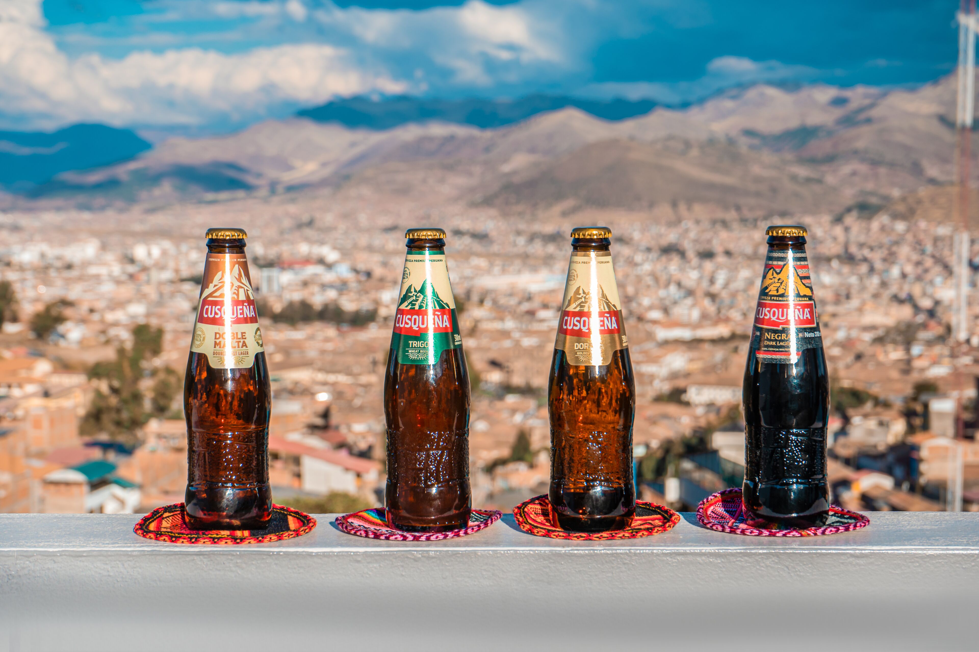 Cuatro botellas de cerveza Cusquena sobre un alféizar, con un paisaje urbano y montañas al fondo.