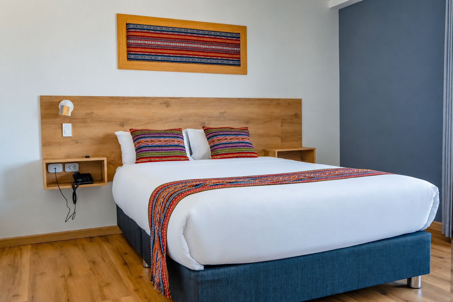 A modern bedroom with a wooden headboard, a bed with white bedding, colorful pillows, a bedside table with a lamp, and a wall-mounted artwork.