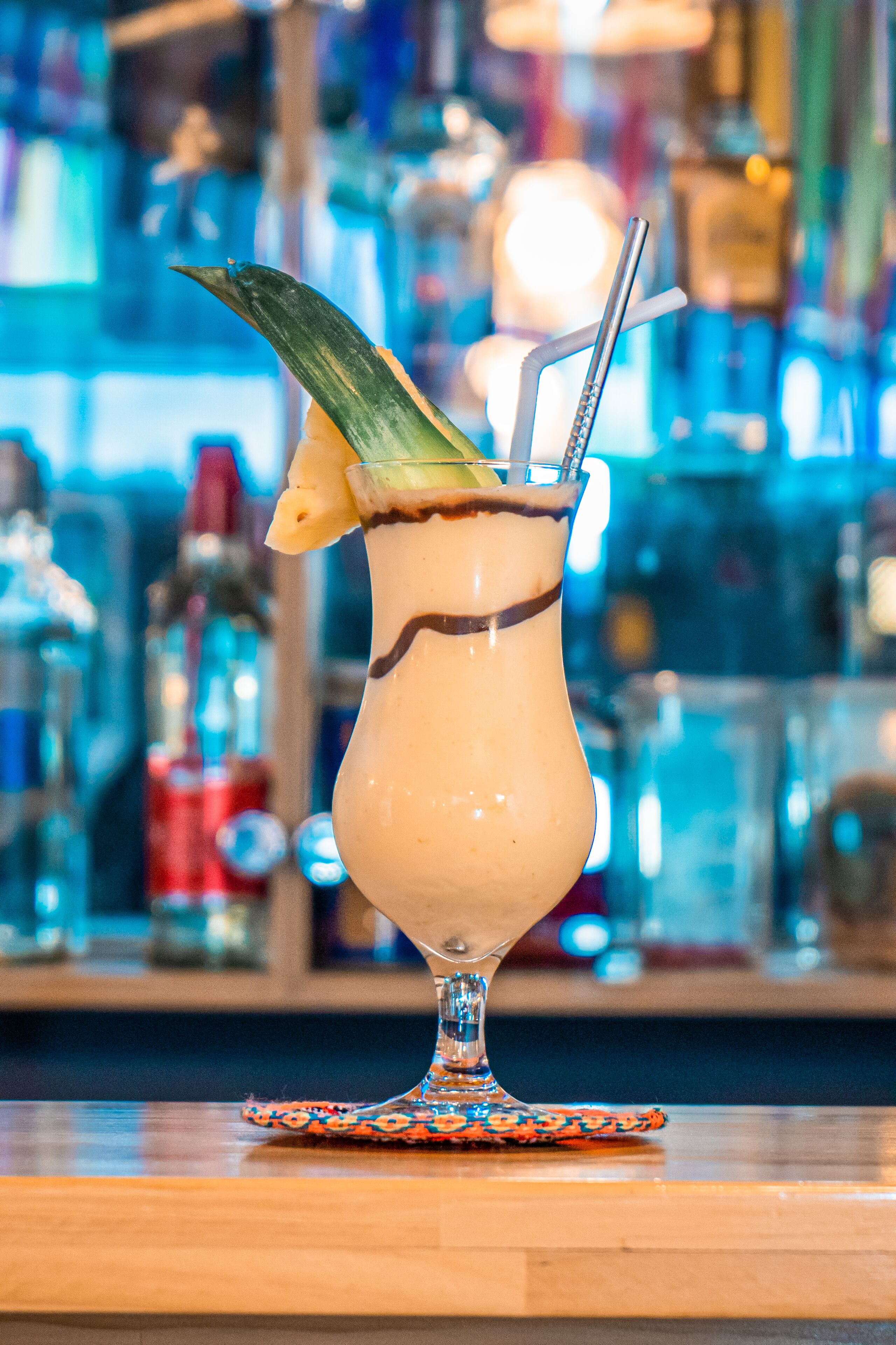 Un cocktail servi dans un verre décoré, agrémenté d’une paille et d’une tranche d’ananas, le tout posé sur un dessous de verre.