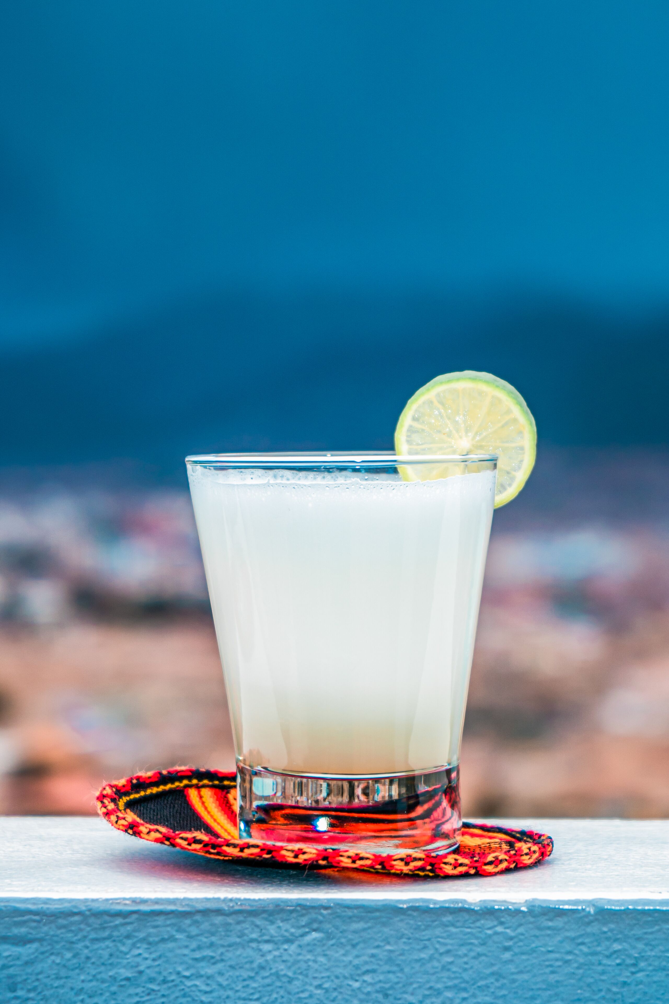 A glass of white beverage with a lime slice on a red and black coaster, placed on a blue surface with a blurred background.