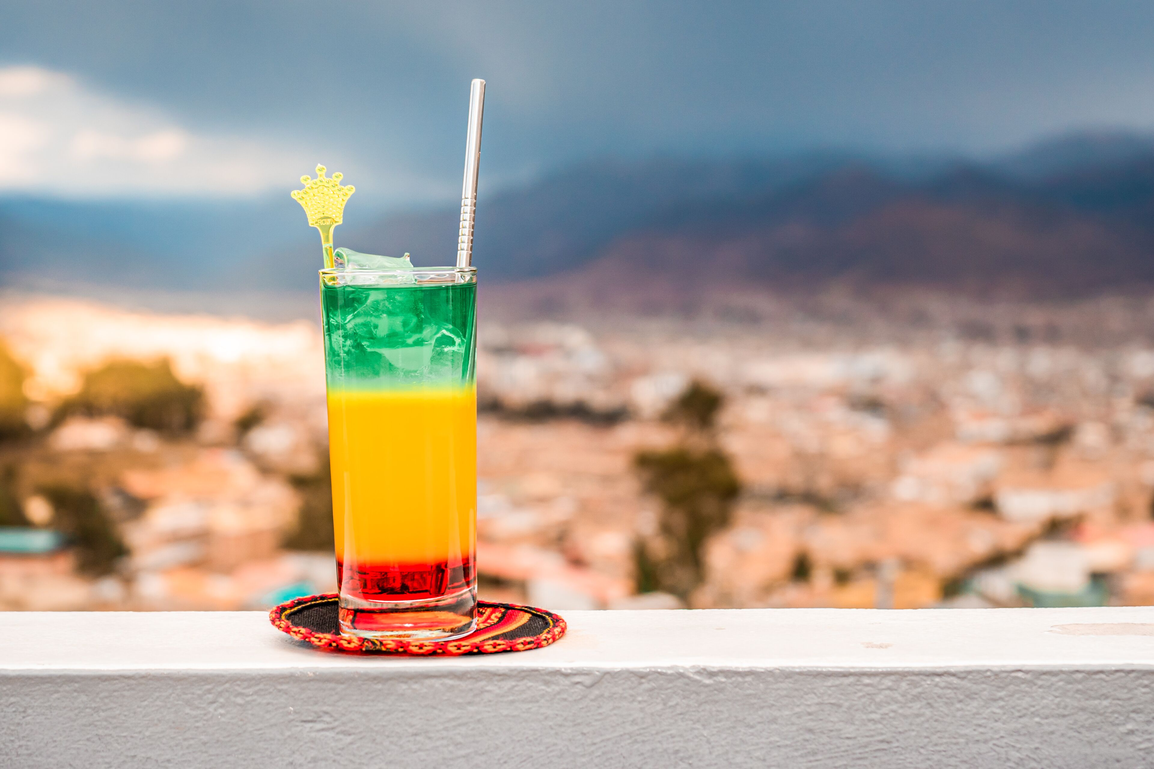 Un vaso con una bebida en capas y una pajita, sobre posavasos, con un paisaje urbano al fondo.