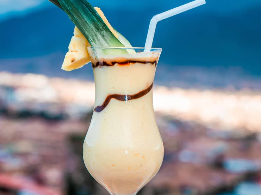 Un cocktail servi dans un grand verre, agrémenté d’une paille, d’une tranche d’ananas et d’une feuille, le tout présenté sur fond de paysage urbain.