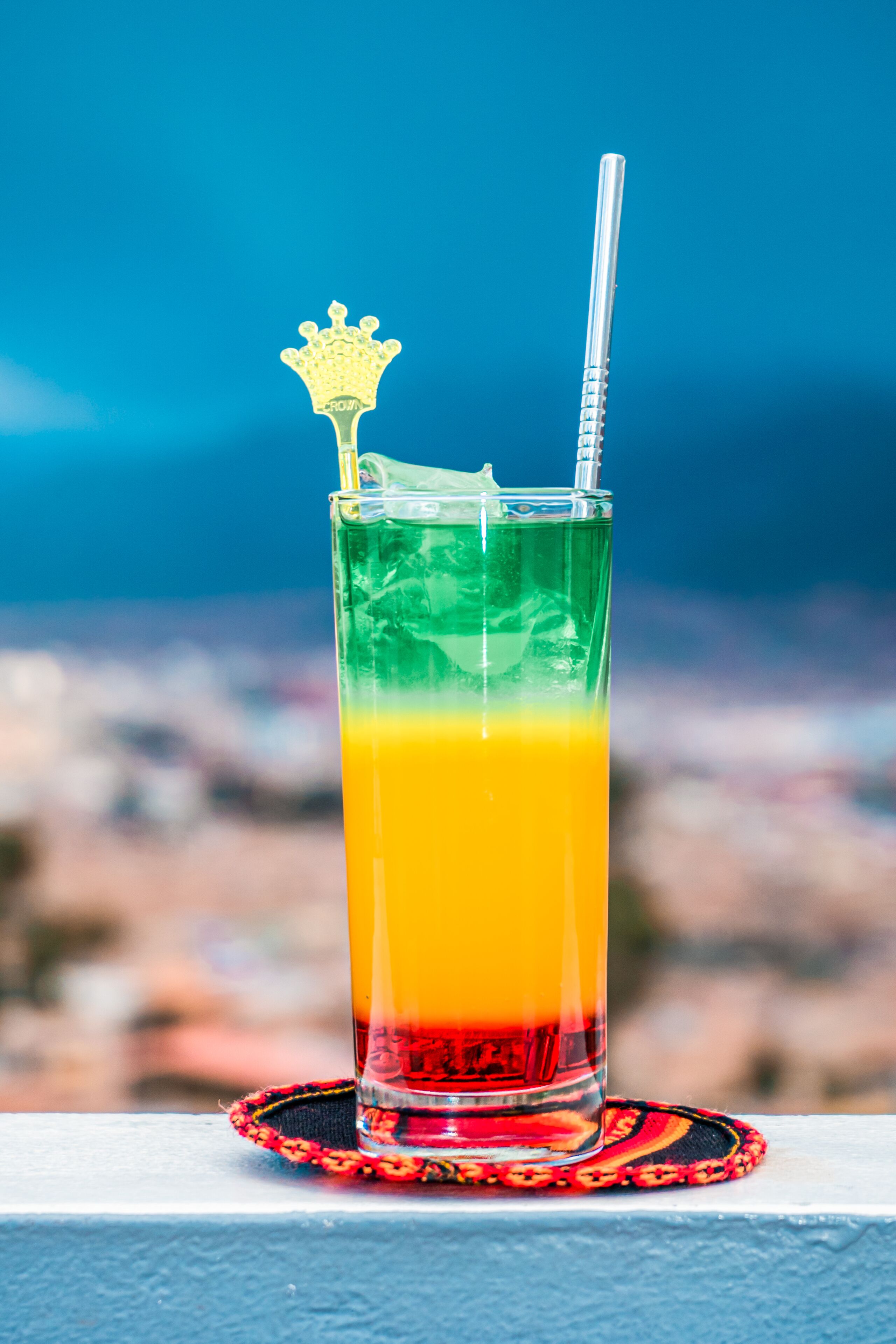 Ett högt glas med en färgövergång från orange till röd vätska, ett sugrör och en dekorativ detalj ovanpå, placerat på en underlägg.