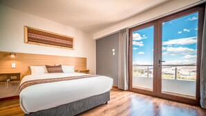 Una habitación de hotel moderna con una cama grande, cabecera de madera, vista a través de una puerta corrediza de vidrio y una obra de arte colgada en la pared.