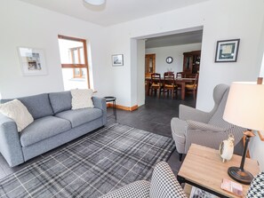 A living room with a grey sofa, a plaid rug, and a houndstooth armchair. There is a dining area in the background with a wooden table and chairs.