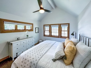 A bedroom with a bed, dresser, windows, ceiling fan, and a bedside lamp.