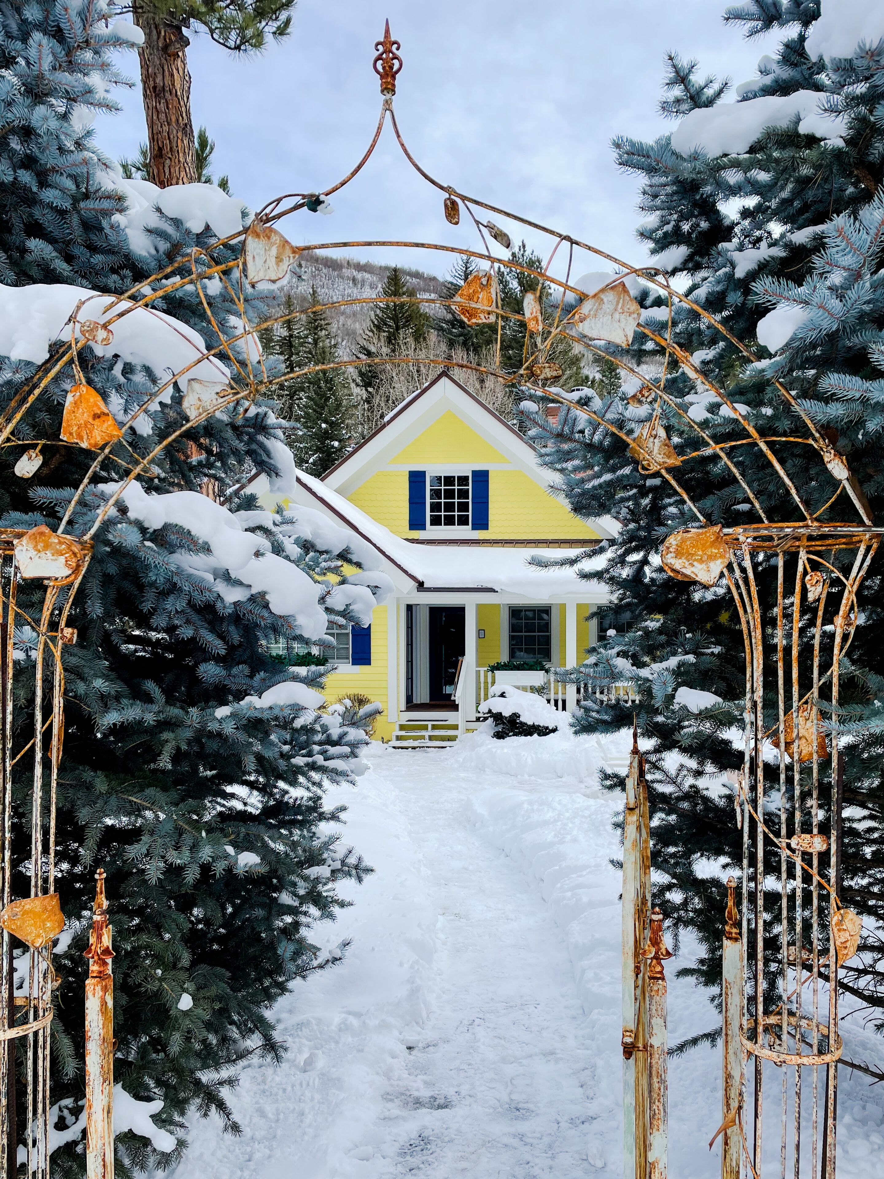 Une maison jaune, avec une porte blanche et des volets bleus, entourée d’arbres couverts de neige et d’une arche métallique ornée de décorations rouillées.