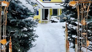 Ett gult hus med en vit dörr och blåa fönsterluckor, omgivet av snötäckta träd och en metallbåge med rostiga dekorationer.