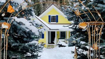 Une maison jaune, avec une porte blanche et des volets bleus, entourée d’arbres couverts de neige et d’une arche métallique ornée de décorations rouillées.