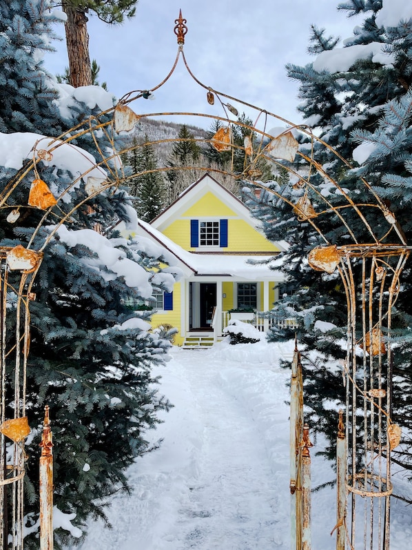 A yellow house with a white door and blue shutters, surrounded by snow-covered trees and a metal archway with rusted decorations.