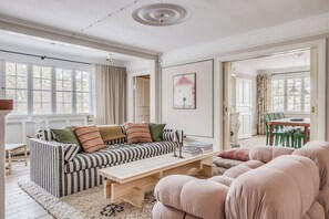 Een woonkamer met een gestreept sofa, een houten salontafel en een roze fauteuil. Een eethoek met een tafel en stoelen is zichtbaar door een open deur.
